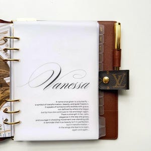 Può includere: Un'agenda in pelle marrone con dettagli dorati, una penna e una chiusura decorativa. L'agenda ha una copertina trasparente con il nome "Vanessa" in elegante calligrafia e una poesia sulla trasformazione.