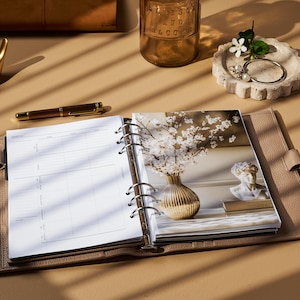 May include: A beige leather planner is open to a blank page, with a photo of white flowers in a vase. A gold pen and a small gold bird figurine are nearby. A small dish with jewelry and a glass bottle are also visible.