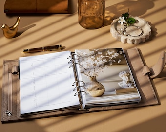 Può includere: Un'agenda in pelle beige è aperta su una pagina vuota, con una foto di fiori bianchi in un vaso. Una penna d'oro e una piccola statuetta di uccello d'oro sono vicine. Sono visibili anche un piattino con gioielli e una bottiglia di vetro.