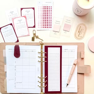 May include: A planner with gold hardware, open to a weekly spread, surrounded by accessories. Includes calendar cards, stickers, a pen, a candle, and a key chain. The planner and accessories are in shades of pink and burgundy.