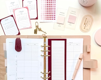 Può includere: Un'agenda con hardware dorato, aperta su una pagina settimanale, circondata da accessori. Include calendari, adesivi, una penna, una candela e un portachiavi. L'agenda e gli accessori sono in tonalità rosa e bordeaux.