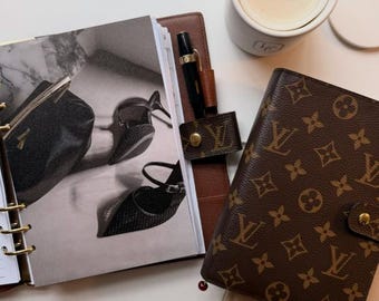 Può includere: Un'agenda aperta con una foto in bianco e nero, un portapenne in pelle marrone con una penna dorata, un portafoglio marrone monogrammato e una candela bianca con uno stoppino nero. Gli oggetti sono disposti su una superficie bianca.