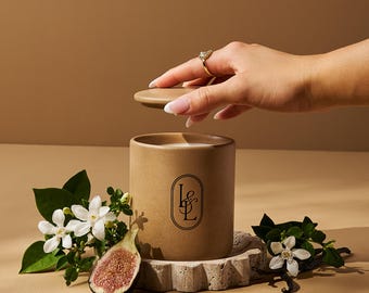 Può includere: Una candela cilindrica marrone con coperchio, con un logo nero, poggia su una base di pietra decorativa. La scena comprende fiori bianchi, un fico tagliato e una mano con un anello d'oro che solleva il coperchio. La candela è circondata dal verde.