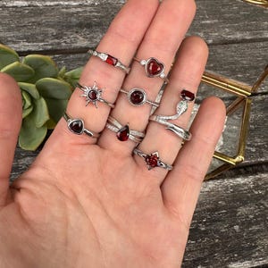 May include: A selection of silver rings, each featuring a different red gemstone design, such as hearts and teardrops. The rings are shown on a hand, with a wooden surface in the background. The gemstones are a deep red colour.