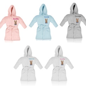 Op de afbeelding: Vijf gepersonaliseerde badjassen voor kinderen met een Peter Rabbit-ontwerp. De badjassen zijn in roze, blauw en grijs. De namen Amelia, George en Cameron zijn geborduurd op de badjassen.