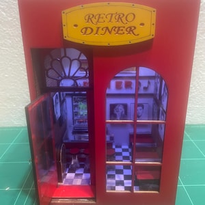 May include: A miniature red Retro Diner model with a yellow sign. The diner has a checkered floor, small tables, and framed pictures on the walls. The open door reveals the interior, and the sign reads "Retro Diner."