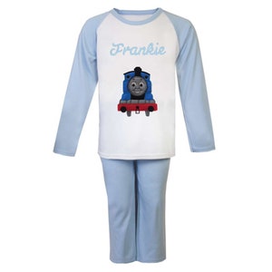May include: Blue and white pajamas with a long-sleeved top and long pants. The top has a blue and white baseball style design with a white front featuring a cartoon image of Thomas the Tank Engine and the name "Frankie" in blue script.