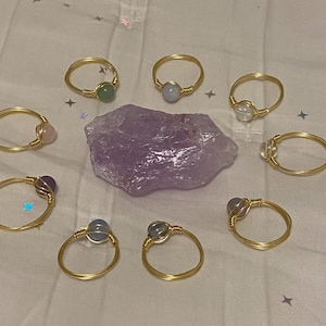 May include: Ten gold wire wrapped rings with different colored gemstone beads. The rings are arranged on a white surface with a purple amethyst crystal in the center.