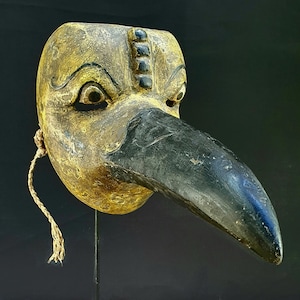 Könnte beinhalten: Eine Holzvogelmaske mit einem langen schwarzen Schnabel, in Gold und Schwarz bemalt. Die Maske steht auf einem schwarzen Metallständer mit quadratischem Sockel.