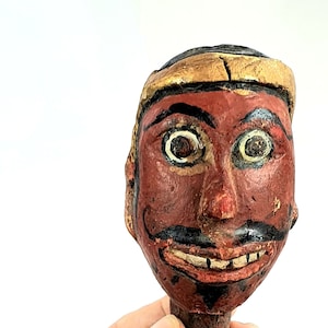 Può includere: Una maschera vintage in legno intagliato a mano con viso rosso, baffi neri e bocca sorridente. La maschera ha una fascia dorata sulla fronte ed è tenuta in mano su sfondo bianco. Un pezzo unico di arte popolare.