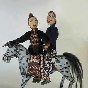 May include: Two wooden puppets, one male and one female, are riding a gray and black spotted wooden horse. The puppets are wearing traditional Indonesian clothing. The horse has a black mane and tail.