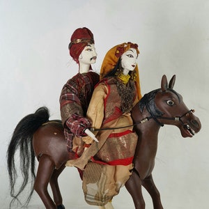 May include: A pair of wooden dolls dressed in traditional Indian clothing riding a brown wooden horse. The male doll is wearing a red and white patterned robe and a red turban. The female doll is wearing a gold and red patterned robe with a yellow scarf. The horse has a black mane and tail.