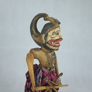 May include: A wooden puppet with a painted face and a long, curved headpiece. The puppet is wearing a colorful skirt and holding two wooden sticks.