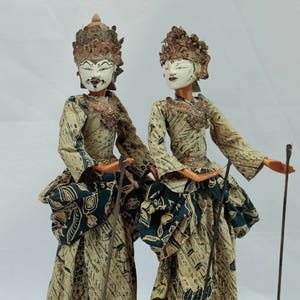 Op de afbeelding: Twee traditionele Indonesische schaduwpoppen, of wayang kulit, met beschilderde gezichten en uitgebreide kostuums. De poppen zijn gemaakt van leer en hout en zijn gekleed in goud en groen gepatroneerd stof.