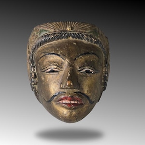 Op de afbeelding: Een houten masker met een gouden gezicht, rode lippen en een zwarte snor. Het masker heeft een kroon met groene en gouden details.