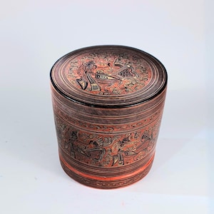 Puede incluir: Una caja de madera redonda y cilíndrica con tapa. La caja está decorada con intrincados diseños pintados en negro y dorado sobre un fondo rojo. Los diseños presentan pájaros estilizados y motivos florales.