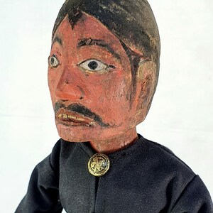 May include: A wooden carved puppet with a painted face, black hair, and a black jacket with gold buttons.