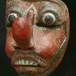 Könnte beinhalten: Eine Holzmaske mit rotem, schwarzem und weißem Farbmuster. Die Maske hat eine große, übertriebene Nase und große Augen. Der Mund ist offen und zeigt Zähne.