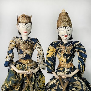 Op de afbeelding: Twee traditionele Indonesische Wayang Golek poppen. De poppen hebben beschilderde witte gezichten met rode lippen, sierlijke gouden hoofdtooien en dragen kleding van blauwe, gouden en groene stof met patronen. Ze zitten op kleine houten standaards.