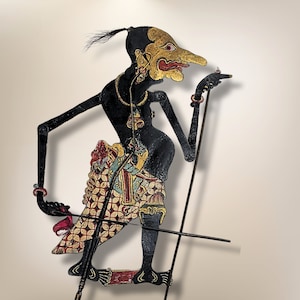 Op de afbeelding: Een zwarte schaduwpop met een lange neus en een uitgebreide hoofdtooi, die een staf vasthoudt. De pop draagt een kleurrijk, gepatroneerd kledingstuk en staat in een staande positie.