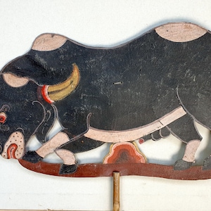 Puede incluir: Una marioneta de sombras negra y blanca de un toro con un ojo rojo y un cuerno dorado. La marioneta está hecha de cuero y está unida a un palo de madera.