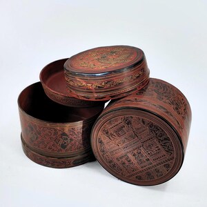 Puede incluir: Tres cajas redondas lacadas en rojo con intrincados diseños en negro y dorado. Las cajas están apiladas una encima de la otra, con la caja superior ligeramente desplazada hacia un lado. Las cajas están hechas de madera y tienen un estilo asiático tradicional.