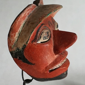 Può includere: Una maschera di legno rossa con un naso grande e una bocca nera dipinta. La maschera ha un cappello dipinto di verde e marrone ed è su un supporto nero.