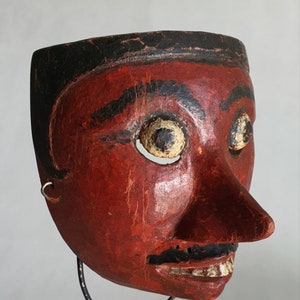 Könnte beinhalten: Eine rote Holzmaske mit schwarzen Details und gelben Augen. Die Maske hat eine große Nase und einen kleinen Mund mit weißen Zähnen. Die Maske steht auf einem schwarzen Metallständer.