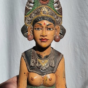 May include: A wooden sculpture of a woman with a colourful headdress and intricate carvings. The sculpture is painted in shades of brown, green, red, and gold.