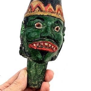 Puede incluir: Una cabeza de títere de madera tallada a mano con una cara verde, labios rojos y cejas negras. El títere lleva un sombrero negro y dorado con una punta puntiaguda.