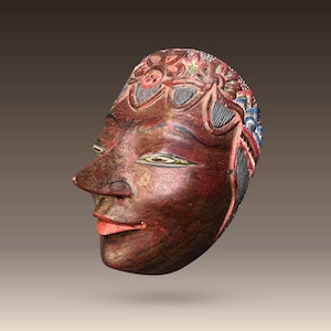 Op de afbeelding: Een houten masker met een gesneden gezicht en ingewikkelde details. Het masker is geschilderd in tinten rood, bruin en goud, met blauwe en witte accenten. Het masker is een traditionele Indonesische kunstvorm.