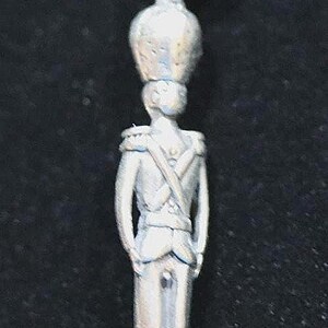 May include: A silver-toned metal charm shaped like a standing soldier wearing a tall bearskin hat and a uniform with cross-body straps. The figure is depicted in a rigid, upright posture with arms at the sides and includes a circular jump ring at the top.