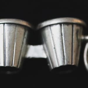 May include: A silver-toned charm in the shape of a pair of bongos. The miniature drums are connected and have a ribbed design. A small loop is attached to the side. The background is black.