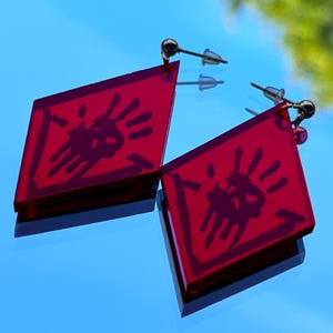Peut inclure: Une paire de boucles d'oreilles rouges en forme de losange. Chaque boucle d'oreille présente un motif d'empreinte de main noire. Les boucles d'oreilles ont des tiges argentées et de petites perles dorées. Elles sont sur une surface bleue réfléchissante.