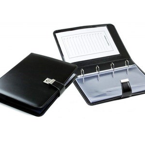 Op de afbeelding: Zwarte lederen map met een slot en een doorzichtig plastic zakje. De map heeft vier ringen en een flap die met een drukknoop sluit.