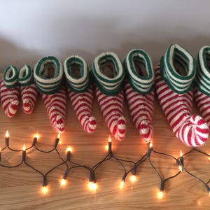 May include: A set of nine red and green striped elf shoes, with the smallest shoe at the left and the largest shoe at the right. The shoes are arranged in a line on a wooden surface with a string of white Christmas lights in front of them.