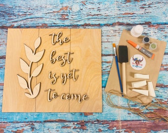 May include: A wooden sign with the text "The best is yet to come" and a leafy branch design. The sign is accompanied by craft supplies including paint, brushes, and foam wedges.