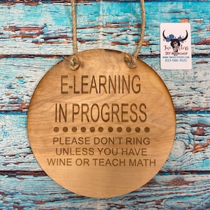 May include: A round wooden sign with the text "E-LEARNING IN PROGRESS PLEASE DON'T RING UNLESS YOU HAVE WINE OR TEACH MATH" engraved on it. The sign is hanging from a rope.