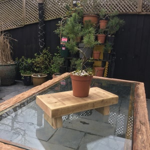 May include: A small pine tree in a terracotta pot sits on a rustic wooden plant stand. The stand is placed on a glass-topped table with a wooden frame.