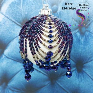 May include: A white Christmas bauble with a beaded design. The beads are in shades of purple, blue, and gold. The bauble is hanging against a blue background.