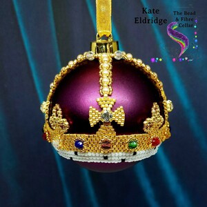 Puede incluir: Una bola de Navidad morada con un diseño de corona dorada con cuentas. La corona está adornada con perlas y piedras preciosas de colores. La bola tiene una cinta dorada para colgar.
