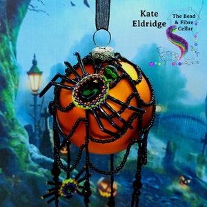 May include: Orange glass Christmas ornament with black beads and a spider web design. The ornament is decorated with green and purple beads and a large green gemstone. The ornament is hanging from a black ribbon.