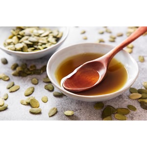 May include: A wooden spoon rests in a bowl of amber liquid, likely oil, with a scattering of green pumpkin seeds. Another bowl of seeds sits to the left. The scene is set on a textured, light gray surface, suggesting a focus on natural ingredients.