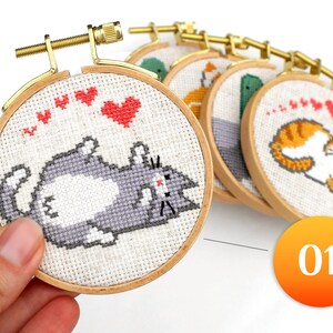 May include: A collection of cross-stitch embroidery hoops featuring cute cat designs. The hoops are round, with wooden frames and gold-colored hardware. The cat designs are in various colors, with red hearts above them. The number "01" is in an orange circle.
