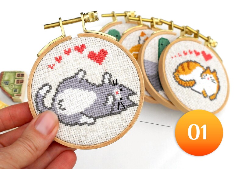 May include: A collection of cross-stitch embroidery hoops featuring cute cat designs. The hoops are round, with wooden frames and gold-colored hardware. The cat designs are in various colors, with red hearts above them. The number "01" is in an orange circle.