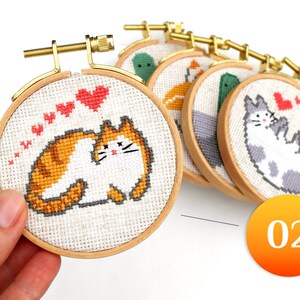 May include: A collection of small, round cross-stitch hoops featuring various cat designs. One hoop is held in a hand, showcasing an orange and white cat with red heart accents. Other hoops display different animal patterns.