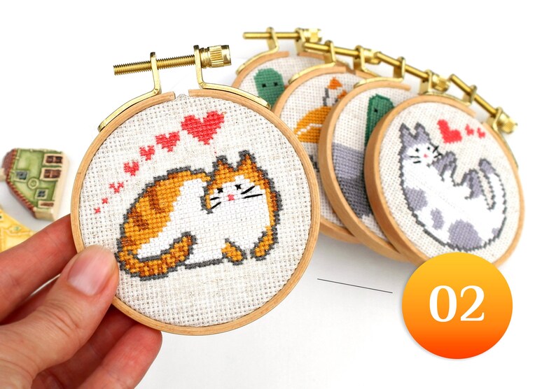 May include: A collection of small, round cross-stitch hoops featuring various cat designs. One hoop is held in a hand, showcasing an orange and white cat with red heart accents. Other hoops display different animal patterns.