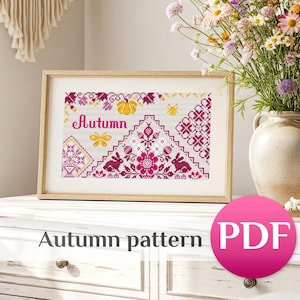 May include: A framed cross-stitch pattern with the word "Autumn" in pink script. The design features pumpkins, flowers, and geometric patterns in shades of pink, yellow, and purple. The image is on a white surface with a vase of flowers to the right.