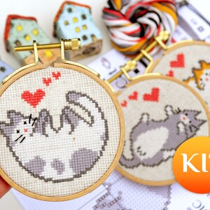 May include: Cross-stitch kit featuring a playful cat design with red hearts. The kit includes embroidery hoops, thread, and instructions. The cat is depicted in gray and white, with a gold-colored hoop. The word "KIT" is in an orange circle.