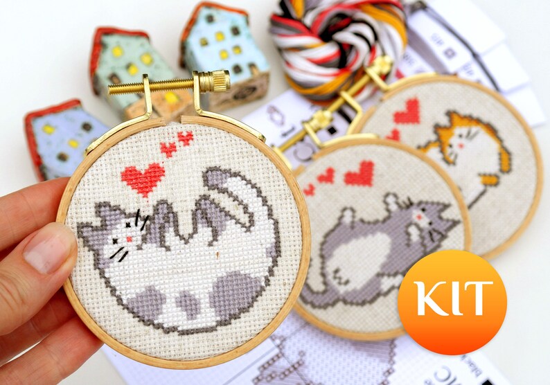 May include: Cross-stitch kit featuring a playful cat design with red hearts. The kit includes embroidery hoops, thread, and instructions. The cat is depicted in gray and white, with a gold-colored hoop. The word "KIT" is in an orange circle.